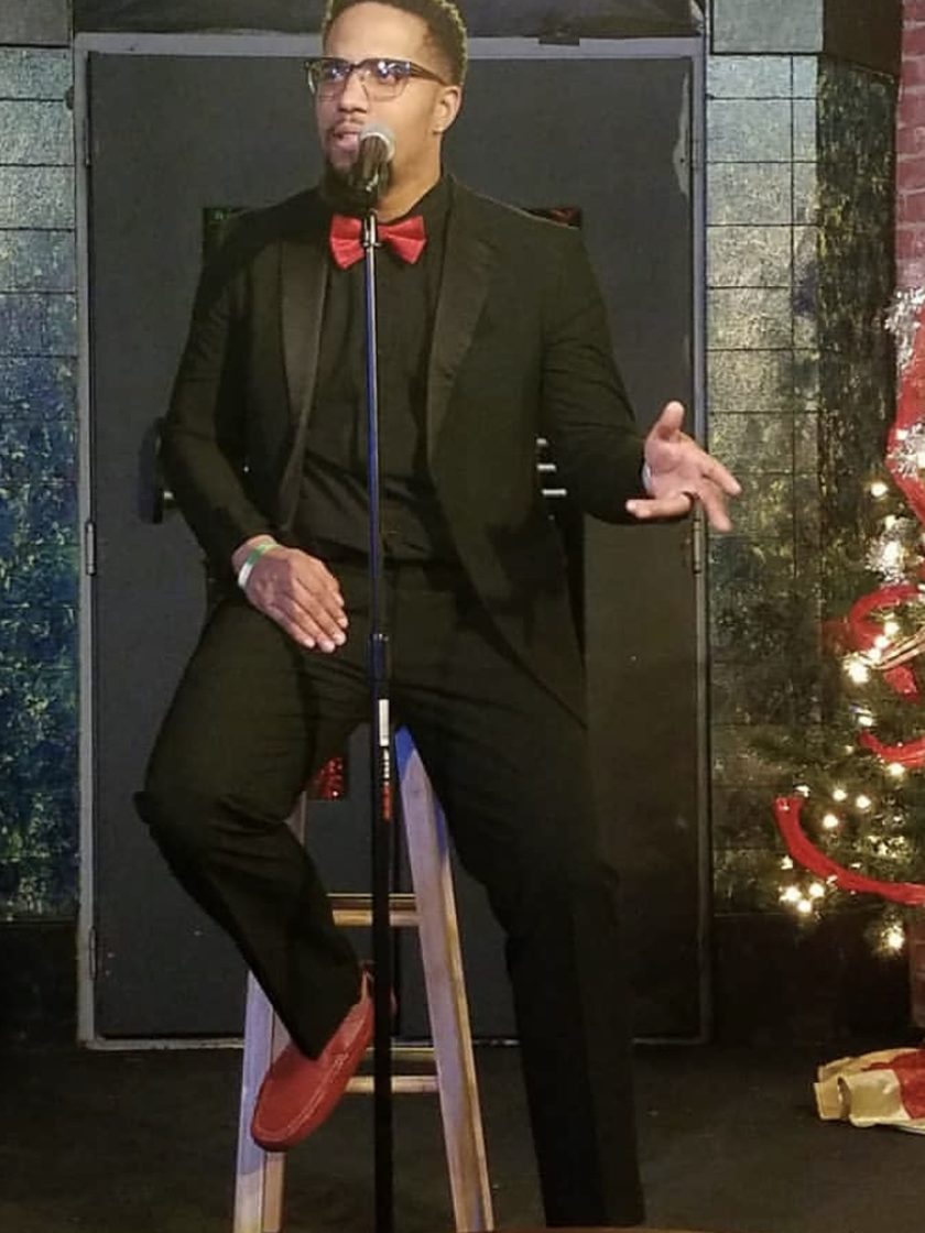 Monterio Thompson seated on a stool in a black suit and red bow tie, one hand open as he talks into a standing microphone.