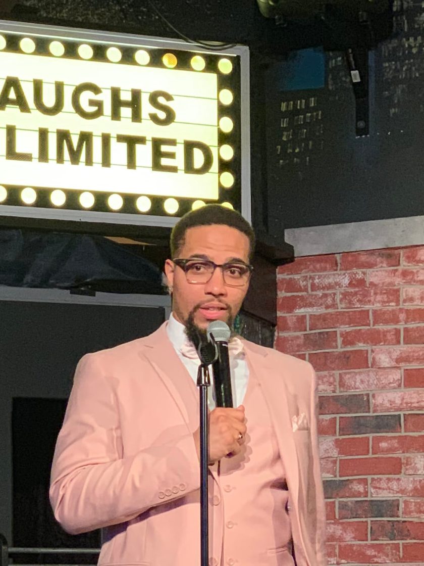 Monterio Thompson hosting in a pink three piece suit, speaking into a microphone under the lit Laughs Unlimited marquee.