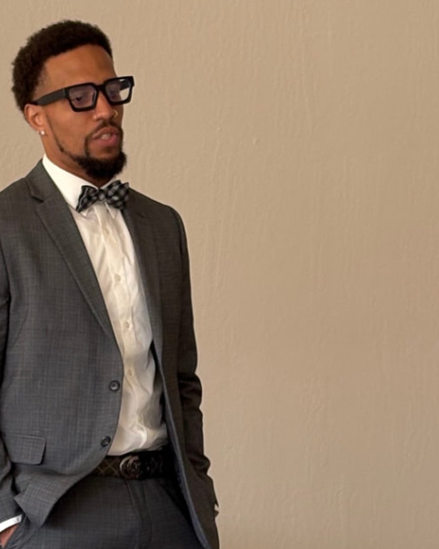 Monterio Thompson, known as Coach Terio, standing in a tailored gray suit with a bow tie and glasses, hands in his pockets, in a community room.