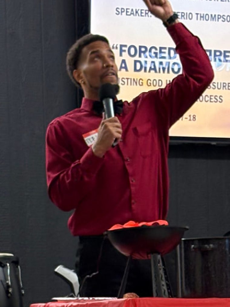Monterio Thompson on stage in a deep red shirt and bow tie, one arm raised high as he speaks into a microphone at a prayer conference.